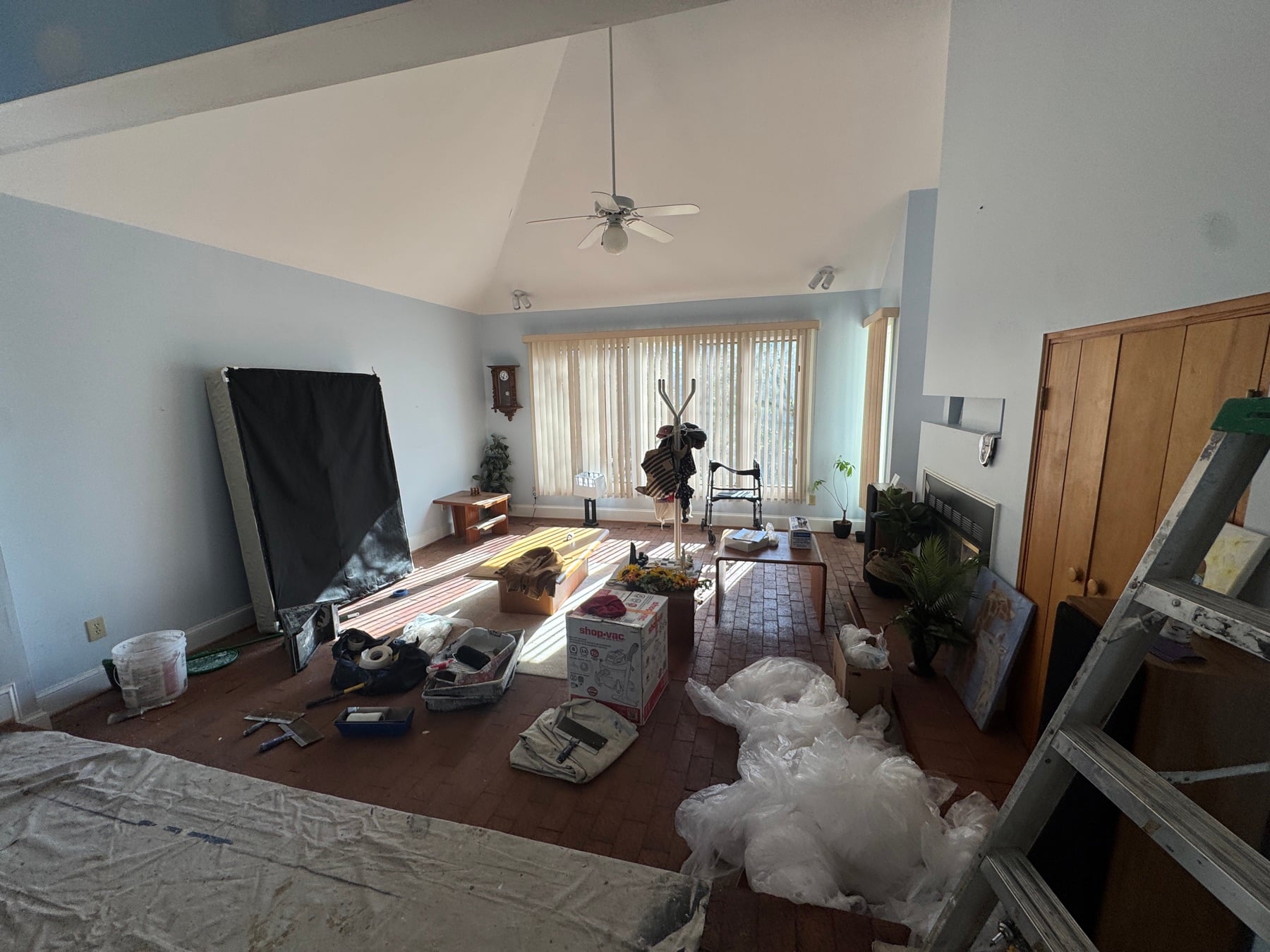 Cluttered living room mid-renovation with drop cloths, ladder and boxes on the brick floor